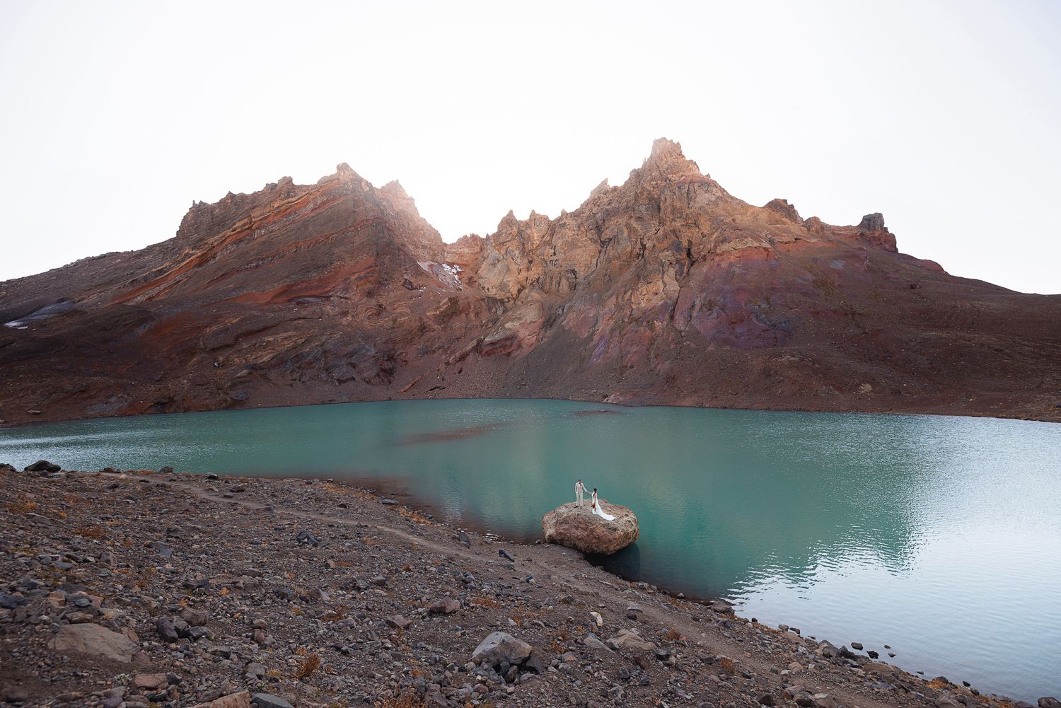 Turquoise alpine lake beneath rugged reddish-brown mountain peaks, with a large boulder resting on the rocky shoreline under a hazy sky.