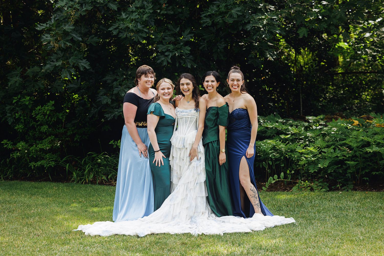 Bride in white gown poses with four bridesmaids in blue and green dresses on a lush green lawn.