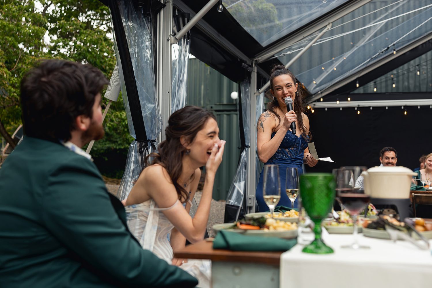 A joyful wedding reception toast with guests laughing as a man speaks into a microphone at an outdoor dinner.