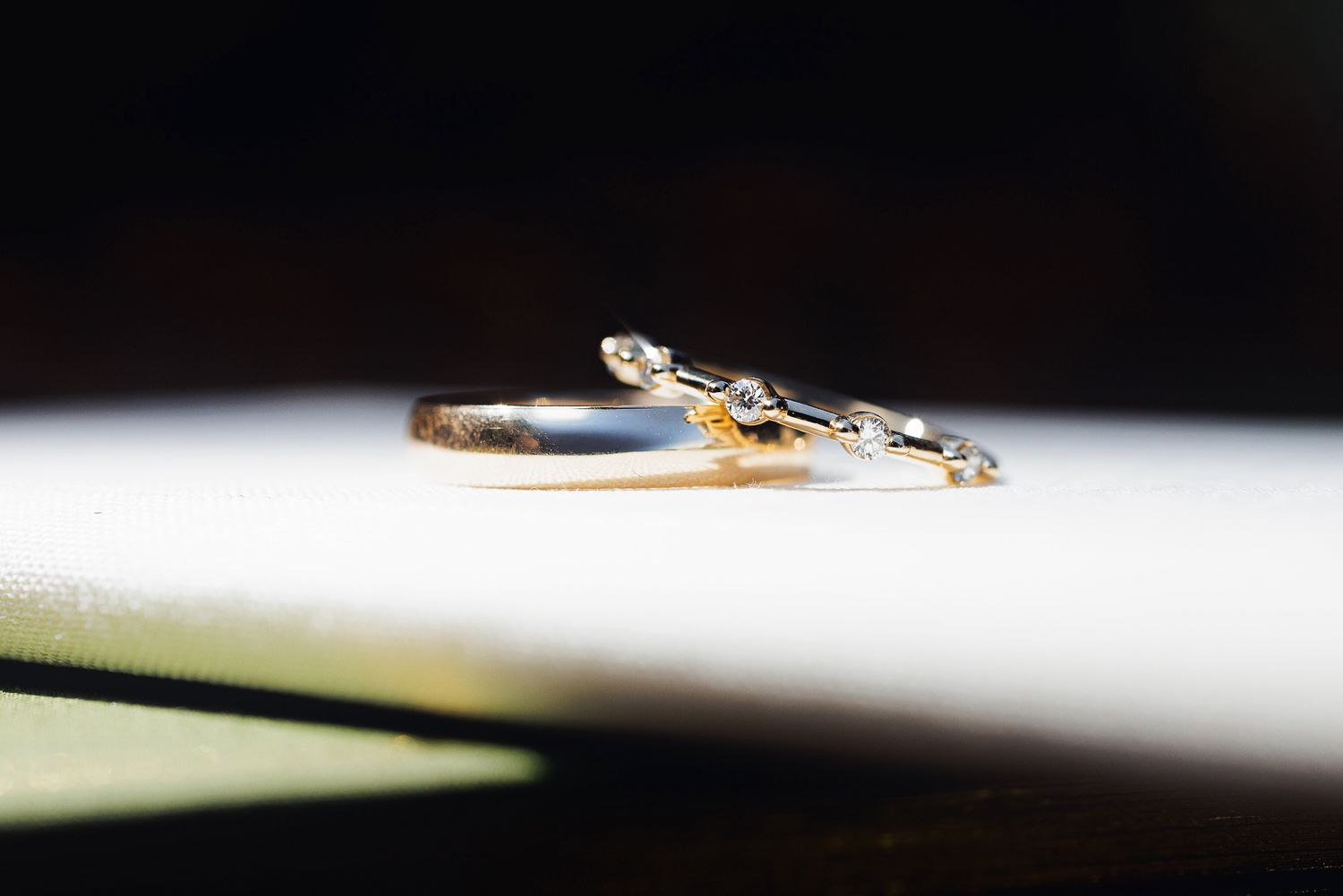 Two delicate gold wedding rings with diamond accents resting on a white surface in soft natural light.