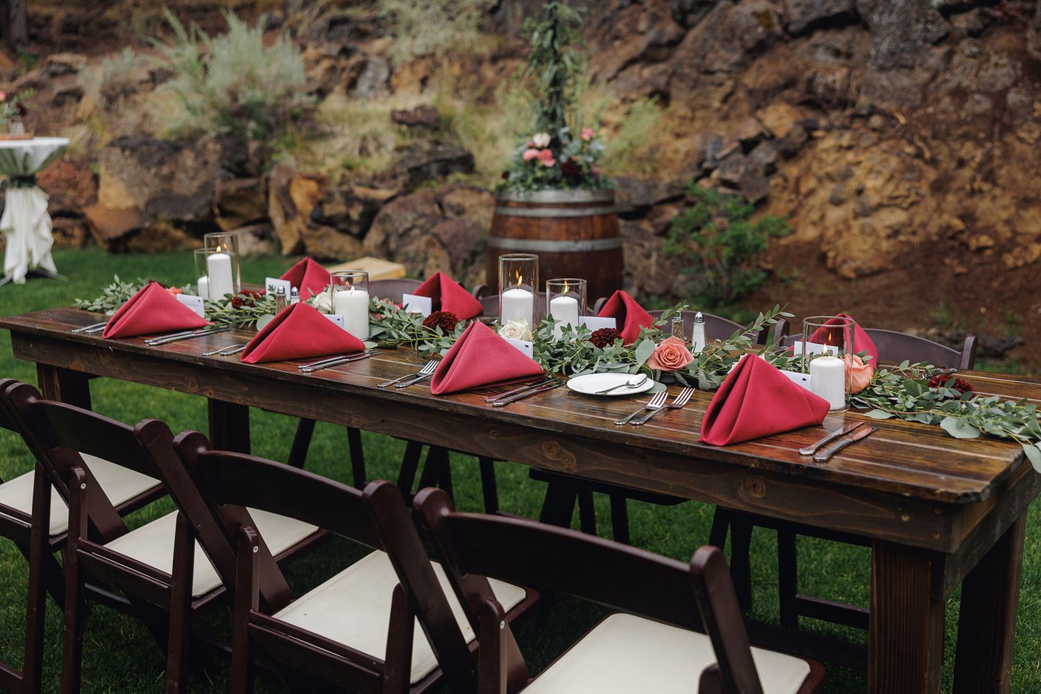 Rustic wooden picnic tables with red umbrellas arranged in an outdoor dining area surrounded by natural scenery.