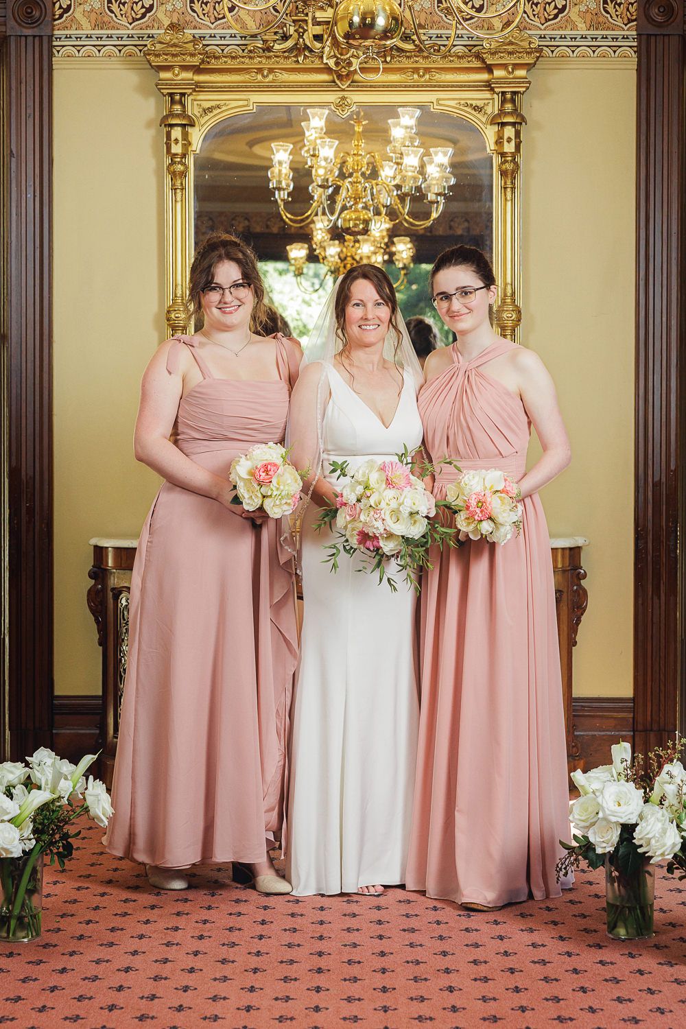 Wedding party poses in an elegant indoor venue with blush pink bridesmaid dresses and floral bouquets.