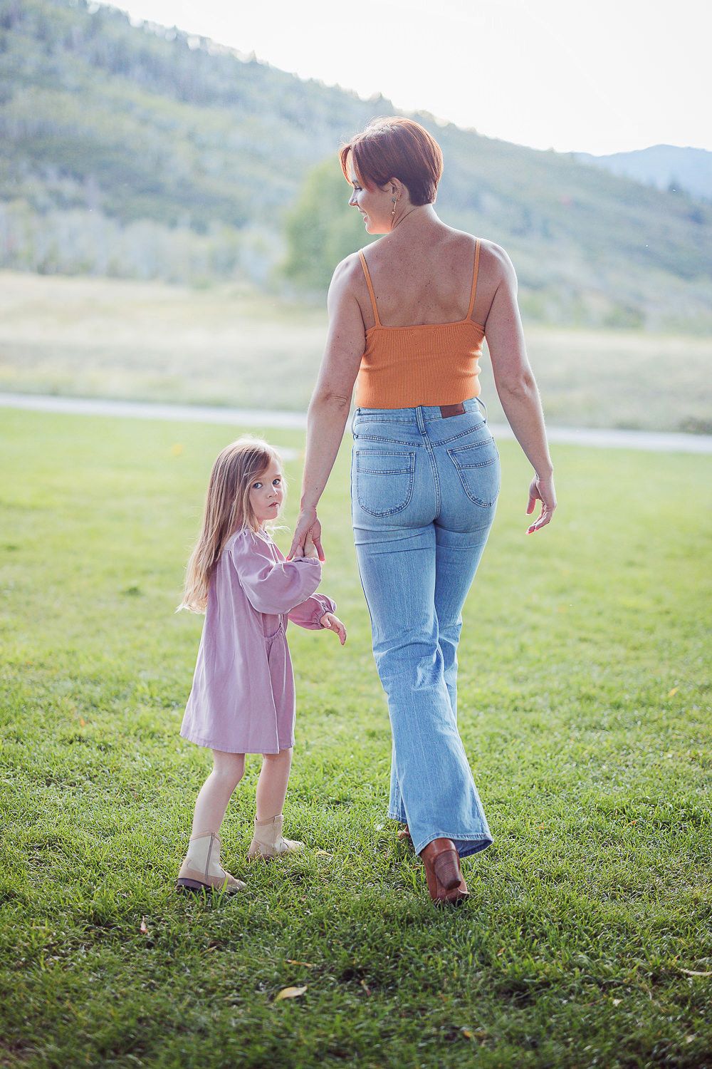 Parent and child holding hands while walking together on grassy hill during golden hour.