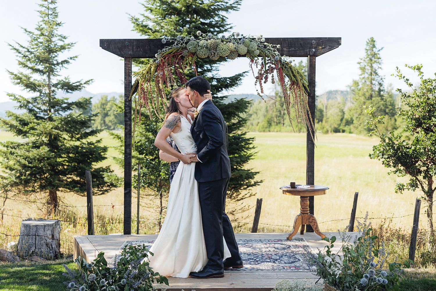 A newlywed couple embraces under a rustic wooden arch surrounded by evergreen trees and mountain scenery.