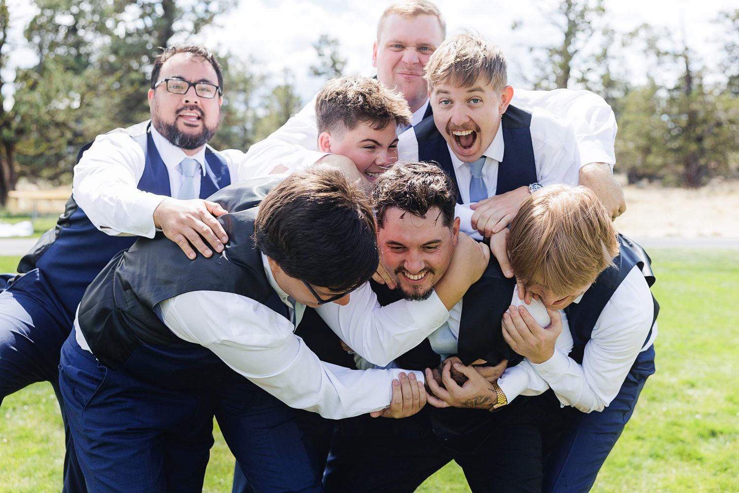 Playful group of groomsmen in navy vests huddling together and laughing during outdoor wedding photos on grass.