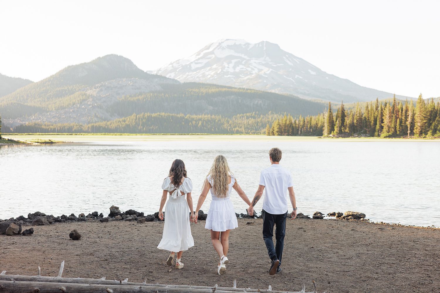 Family of three holding hands walking toward a serene mountain lake surrounded by pine trees and snow-capped peaks.