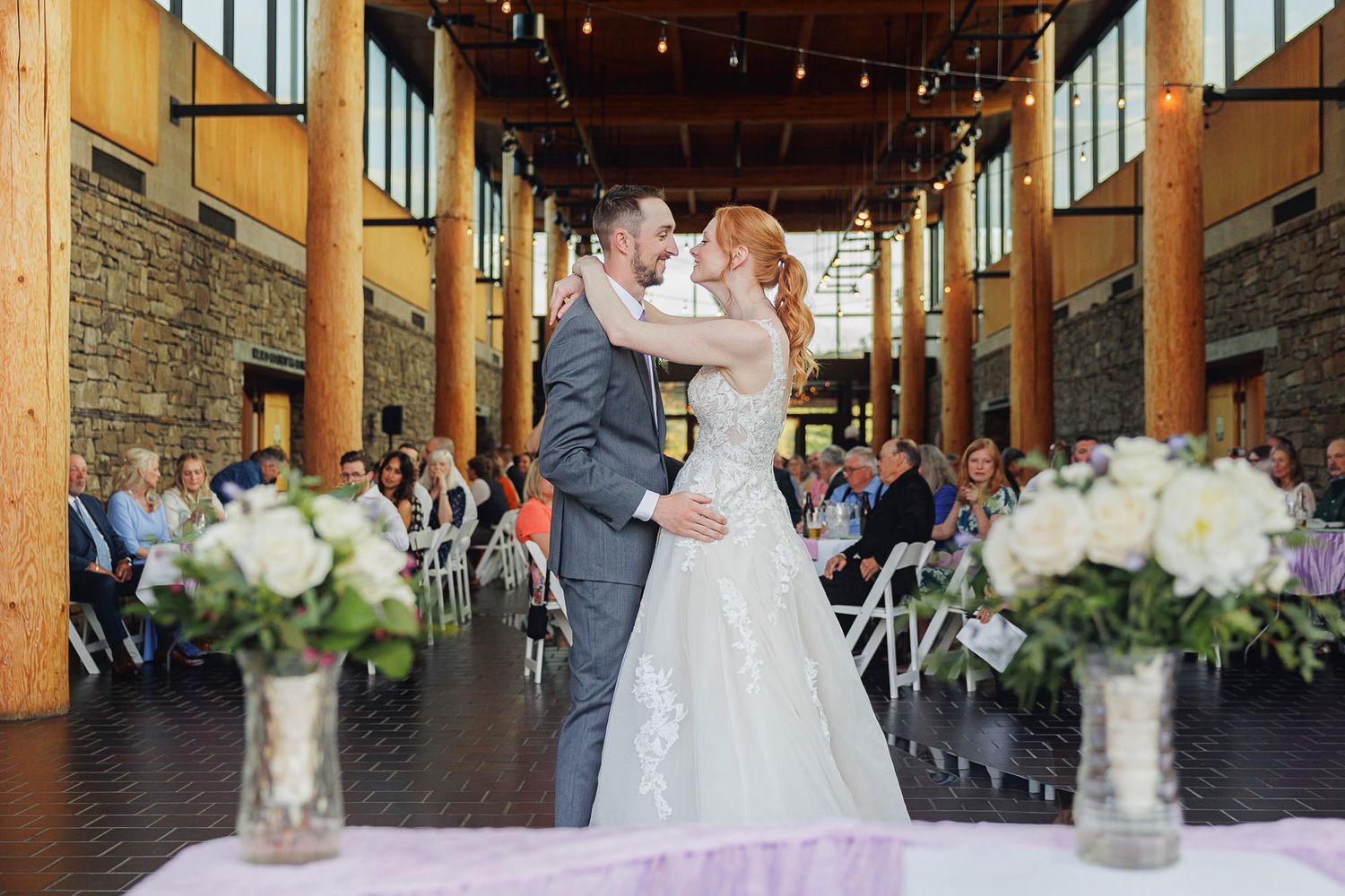 Bride and groom share first kiss at indoor wedding ceremony with stone walls, floral arrangements, and seated guests.