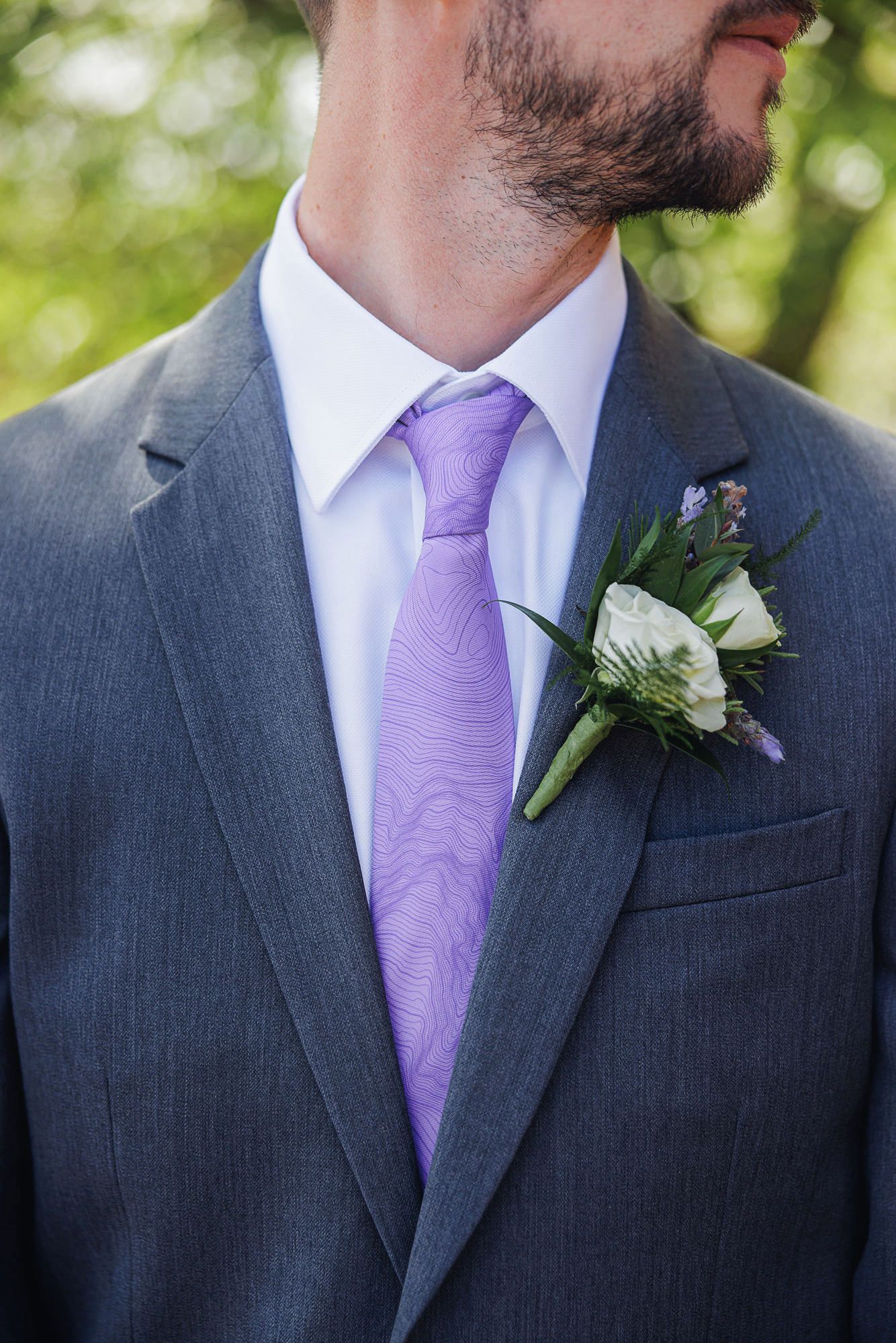 Groom in gray suit with lavender tie and white floral boutonniere with purple accents at a wedding.