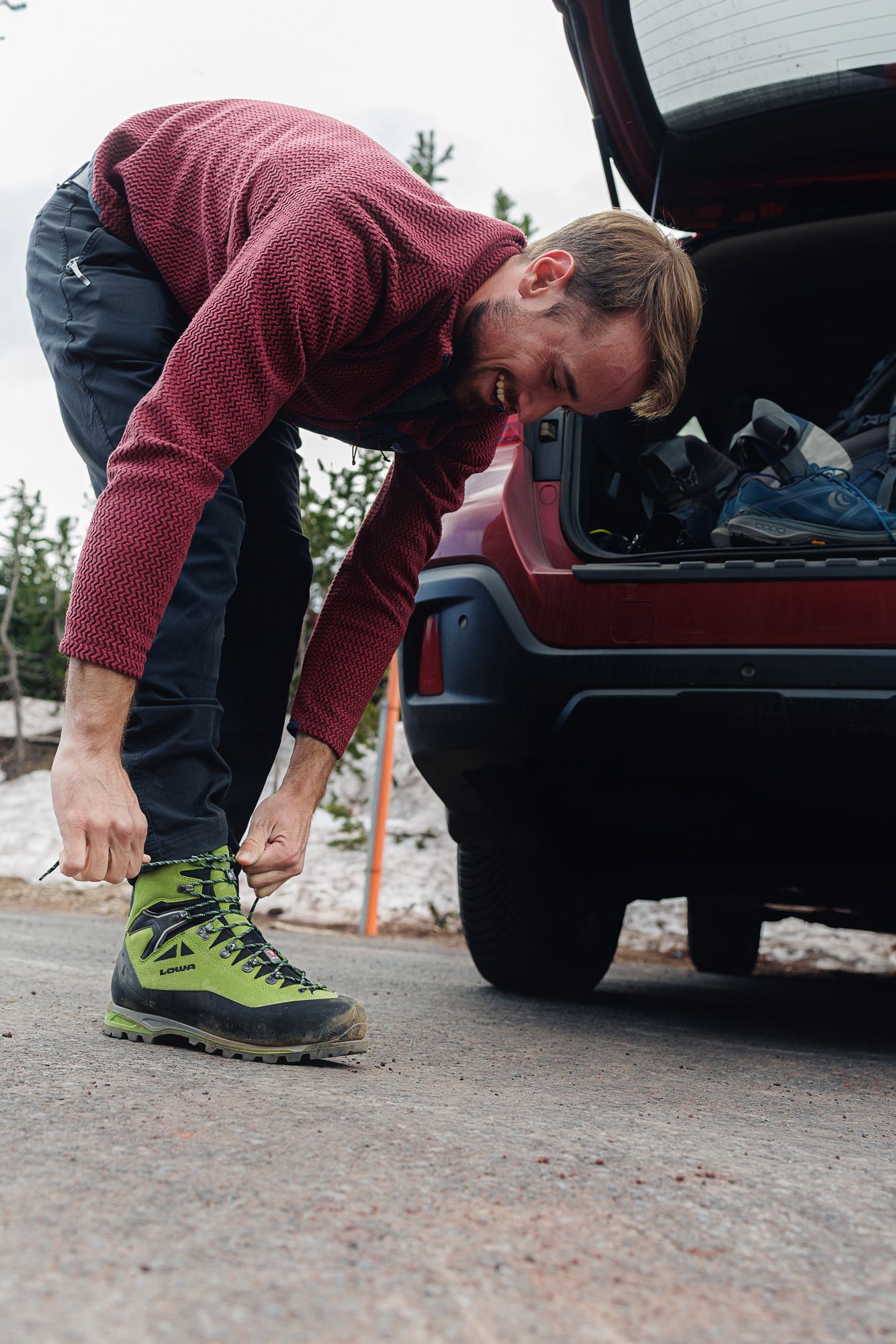 Man in red sweater tying green hiking boots at car trunk outdoors.