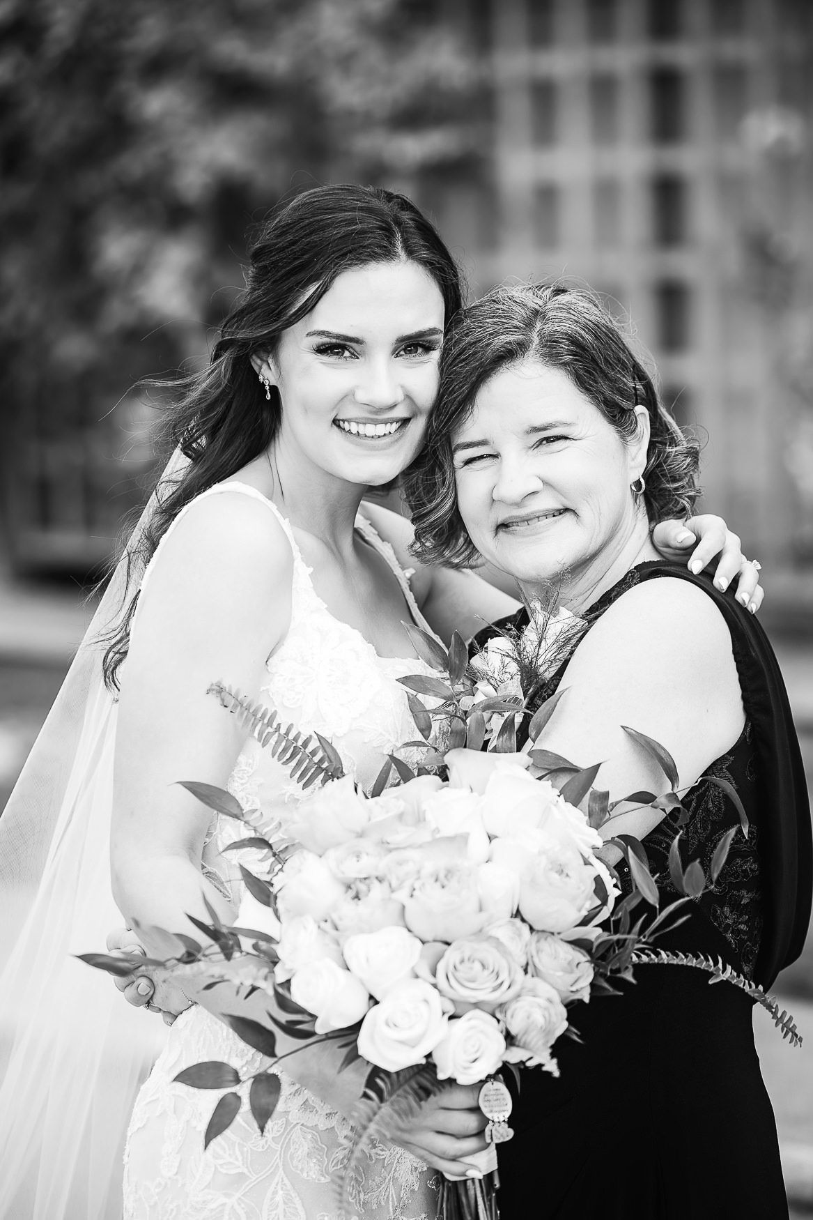 Two women pose together at a wedding celebration, with one wearing a white bridal gown holding a floral bouquet.