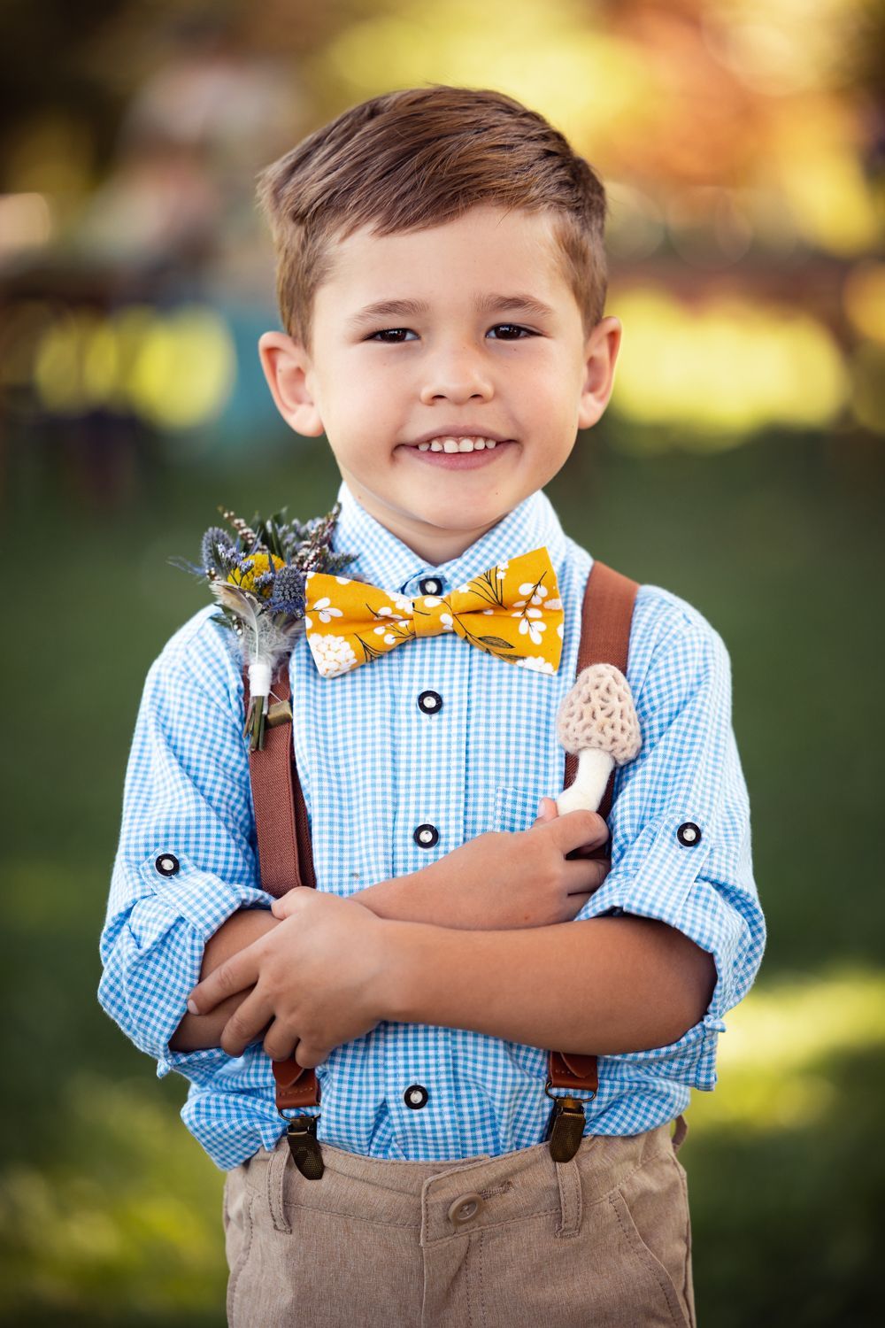 Young child wearing blue gingham shirt with yellow bowtie and brown suspenders smiling outdoors.