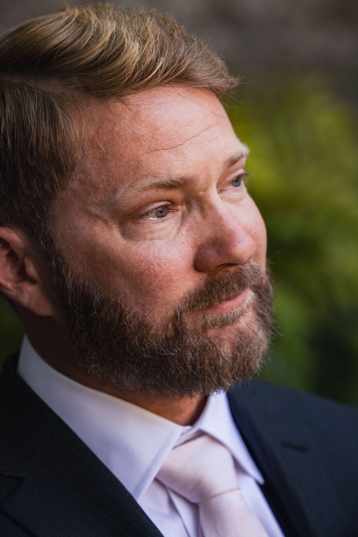 Close-up portrait of formally dressed person with full beard wearing navy suit and pink tie looking to side outdoors.