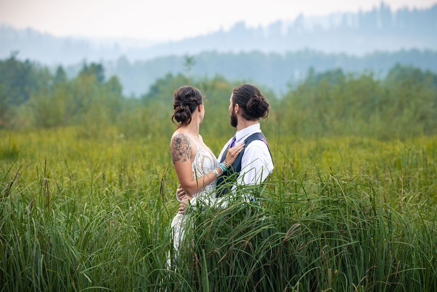 A romantic wedding couple stands together in a misty meadow with tall grass and forest mountains in the background.