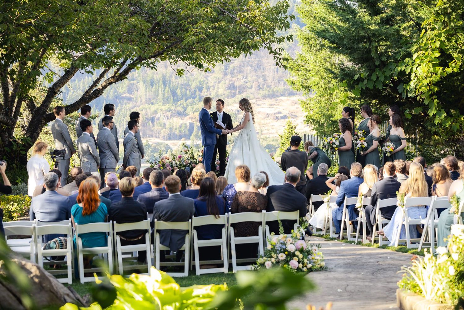 A scenic outdoor wedding ceremony in a garden with white chairs surrounded by lush trees and mountain views.