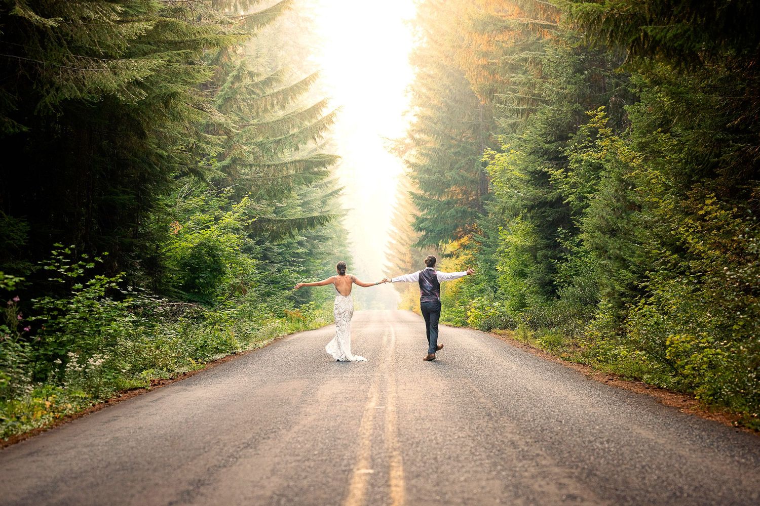 A couple walks down a sunlit forest road surrounded by evergreen trees.