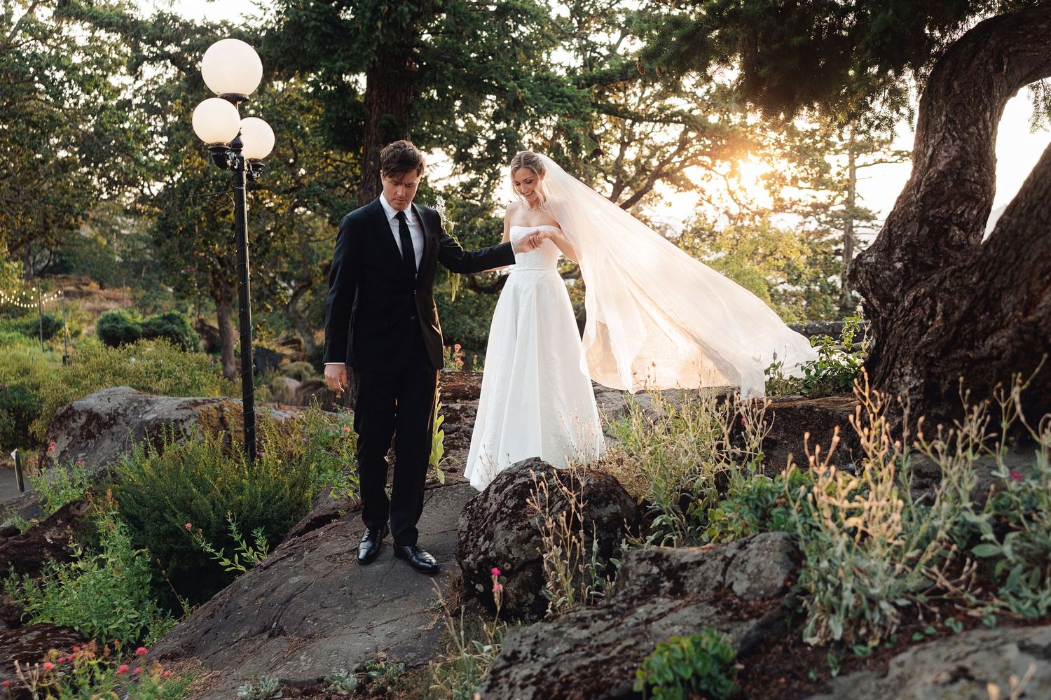 Bride and groom walk hand-in-hand outdoors at sunset, her veil flowing in the breeze among lush garden greenery.