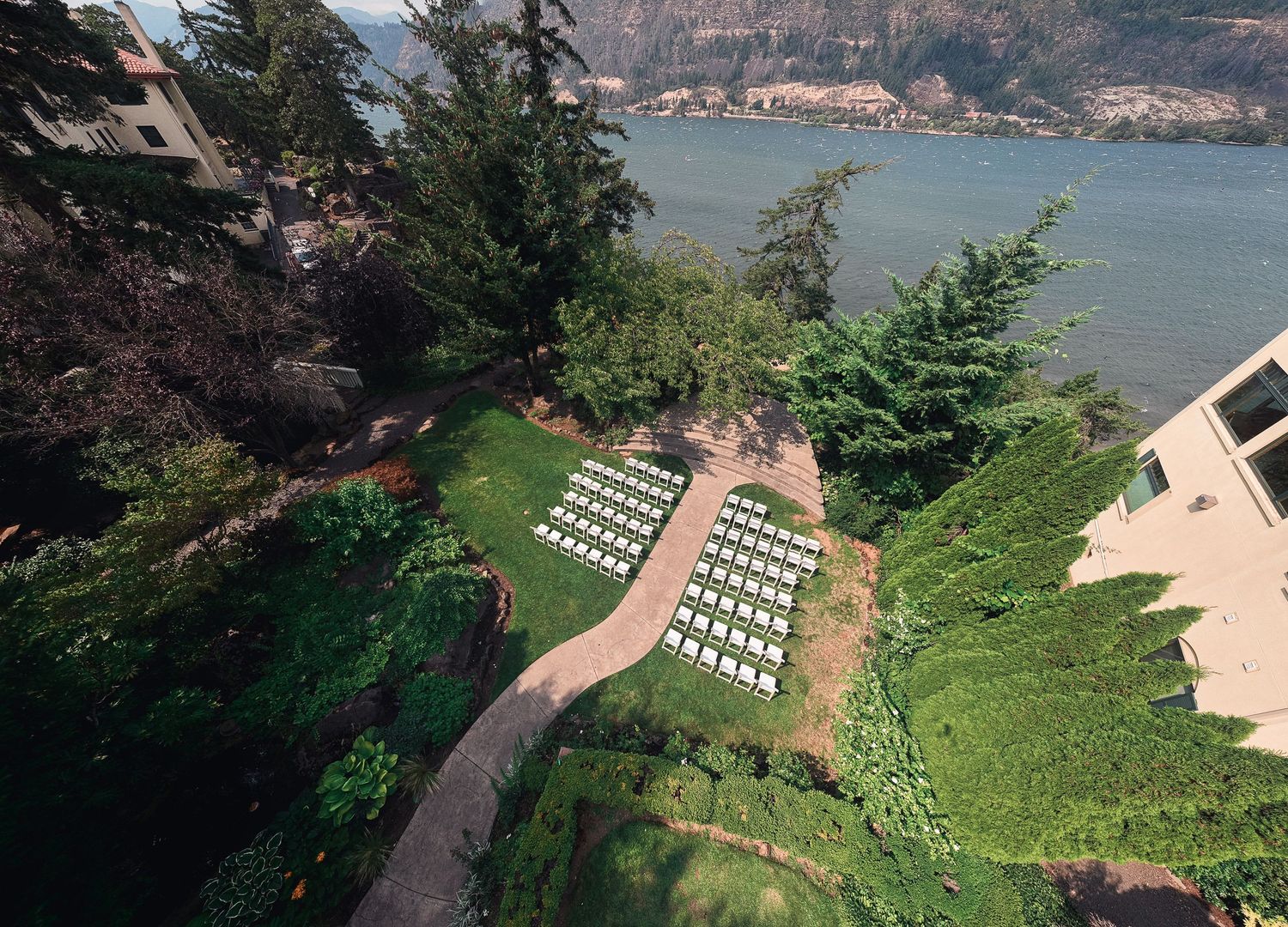 Aerial view of white chairs arranged for an outdoor wedding ceremony on a lush green hillside overlooking a scenic river.