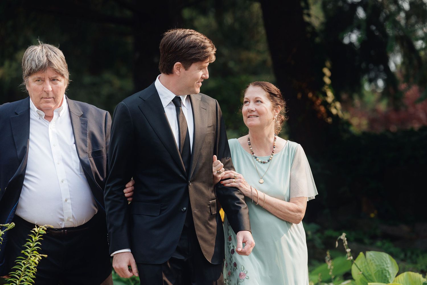 Elegant family trio walking outdoors in formal attire, young man in suit flanked by older man and woman in garden setting.