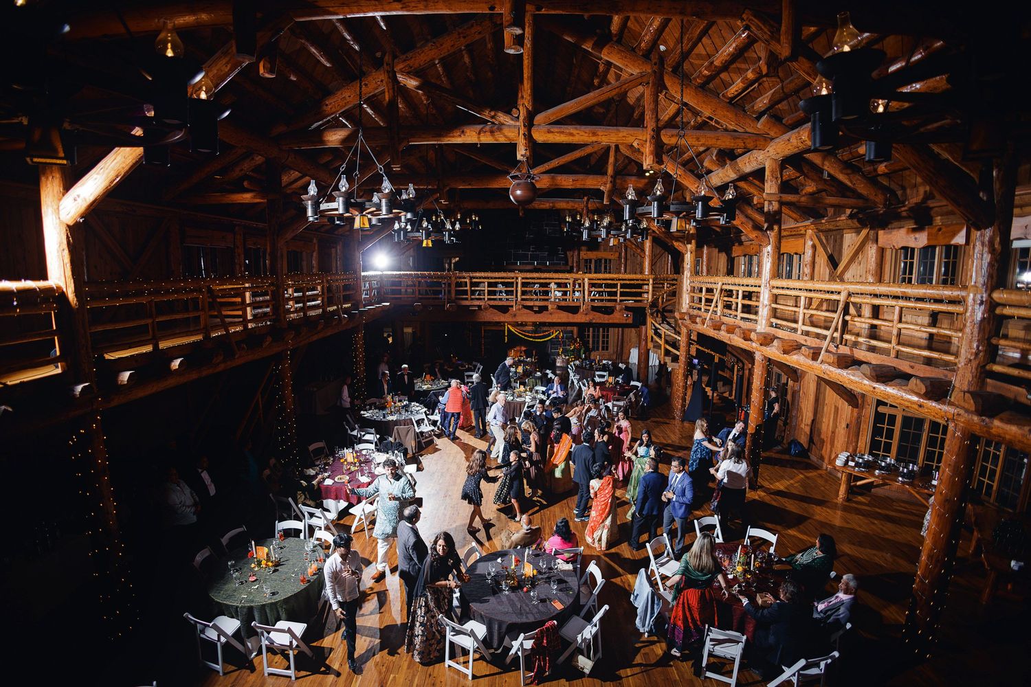 Aerial view of a rustic barn wedding reception with wooden beams, round tables, and guests mingling on the dance floor.