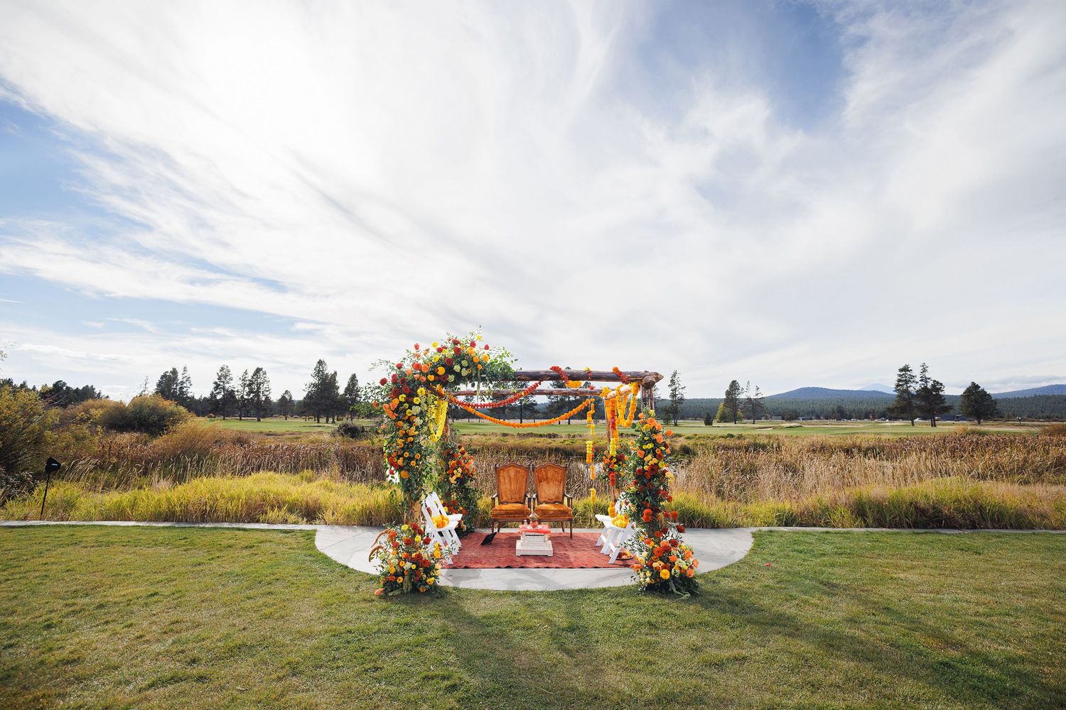 Colorful floral mandap wedding ceremony setup on green lawn with mountain backdrop under blue sky.