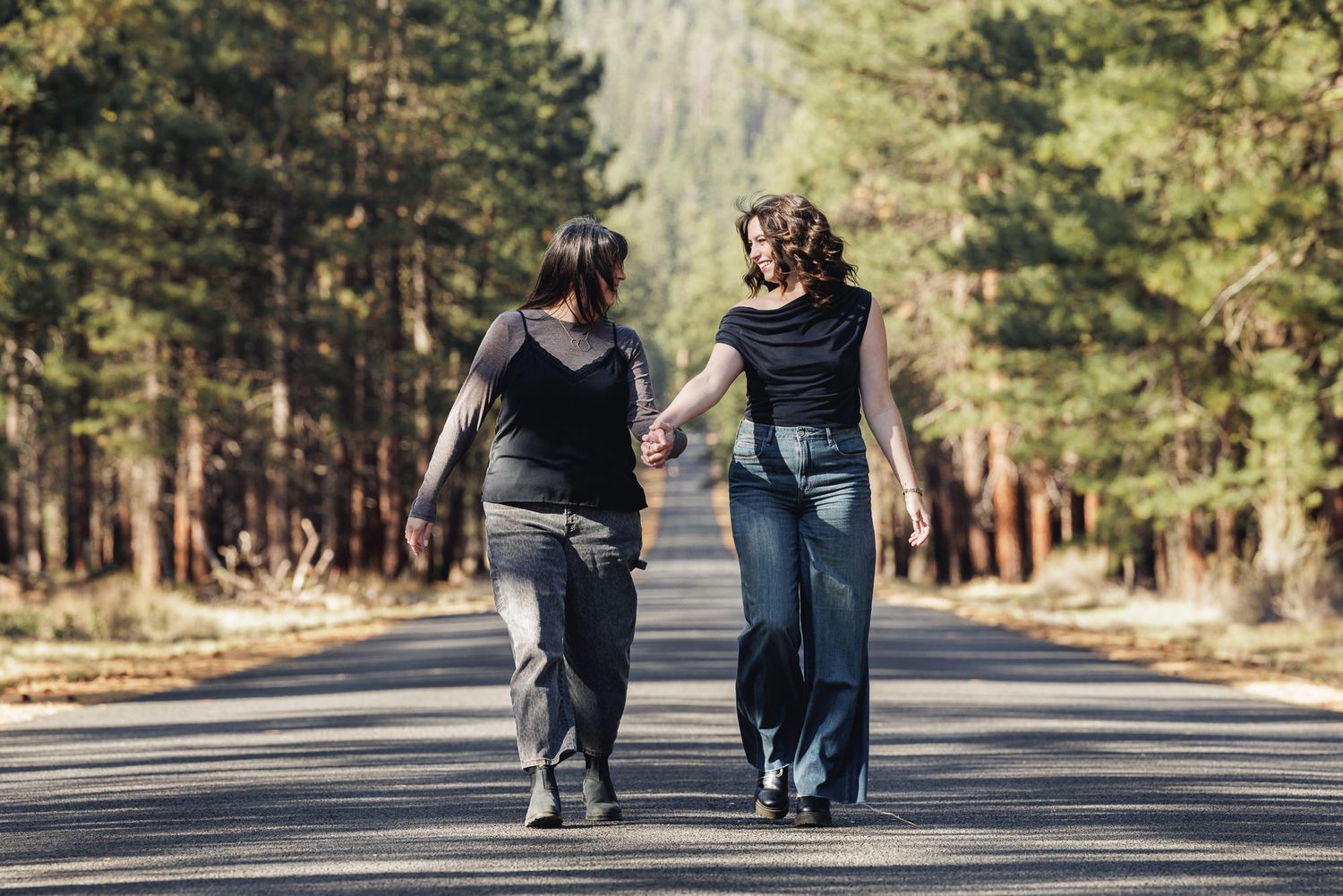 Two women walk hand-in-hand down a tree-lined road, viewed from behind, wearing casual dark outfits.