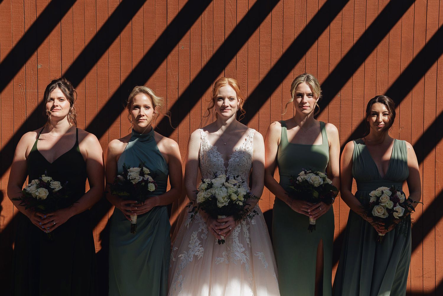 Bride in lace gown with four bridesmaids in sage green dresses holding bouquets, posing before a rustic red barn wall.
