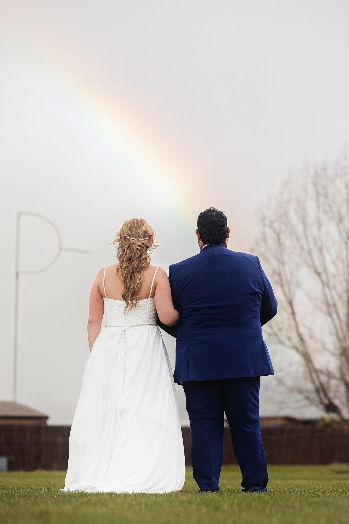 Bride and groom standing together viewing a rainbow on their wedding day, seen from behind.