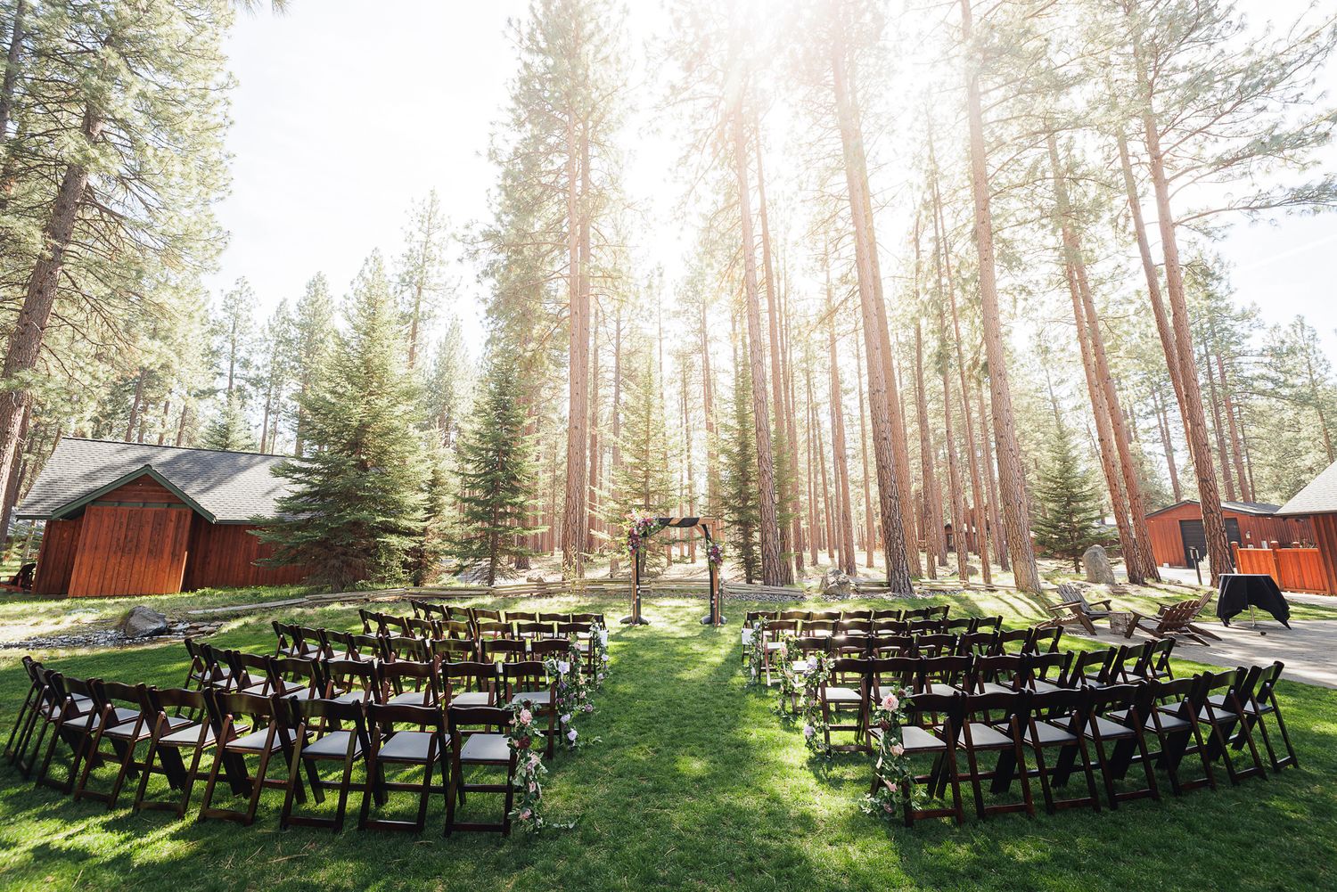 Outdoor wedding ceremony setup with rows of dark chairs on green grass surrounded by tall pine trees and rustic red barn.