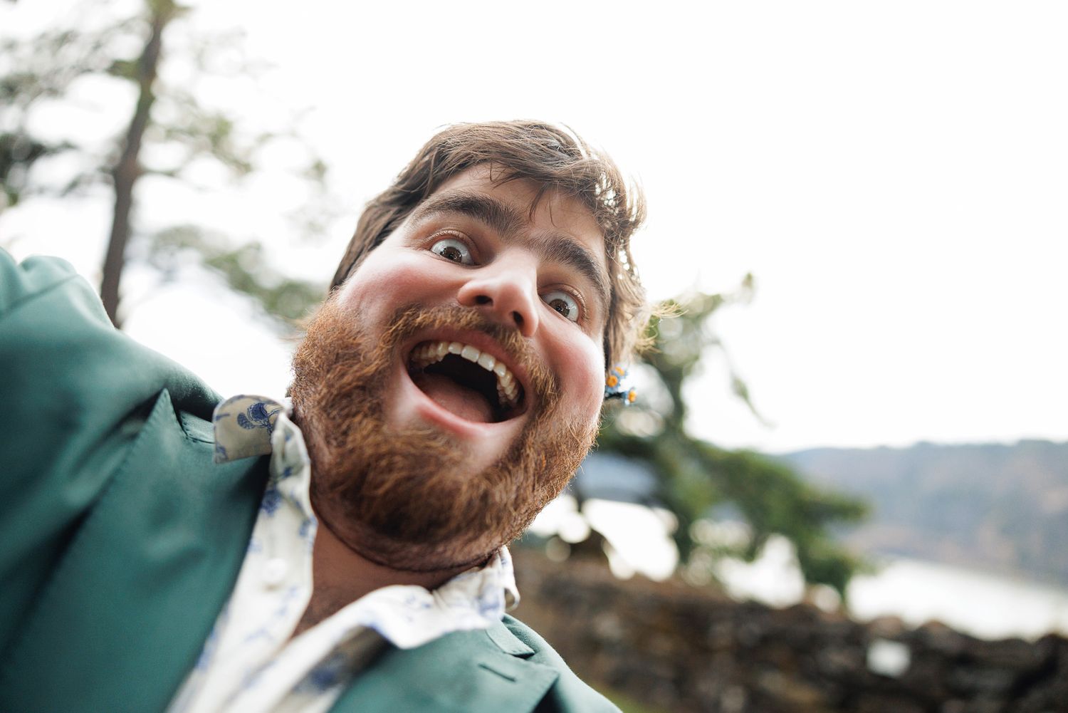 Excited bearded man in green jacket taking a wide-angle selfie outdoors with trees and rocks in background.