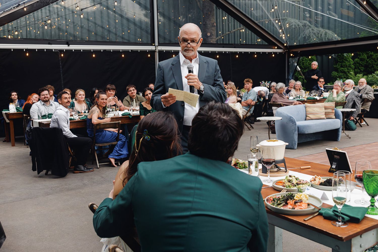 Man giving a speech at an elegant outdoor wedding reception dinner under a glass marquee tent.