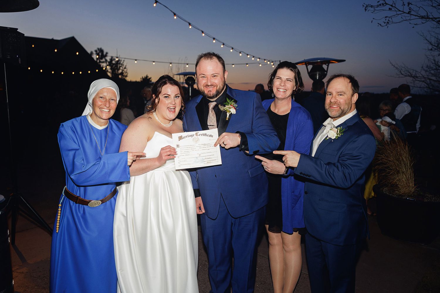 People in coordinated blue and white outfits gather for an outdoor evening celebration with string lights in the background.