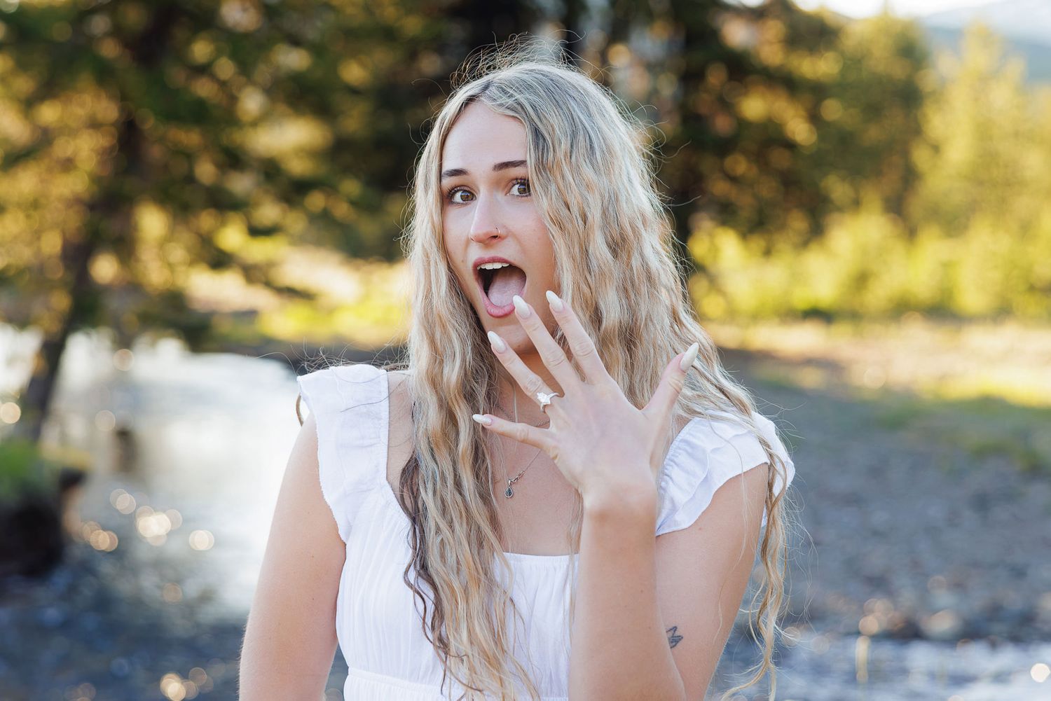 Surprised blonde woman in white dress with hand raised near face outdoors near trees.