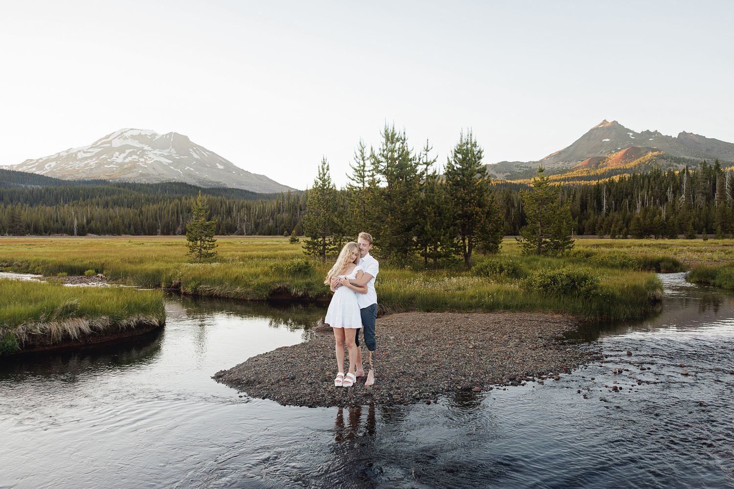 Couple embracing on a rocky stream bank with mountain peaks and pine trees in the background at golden hour.