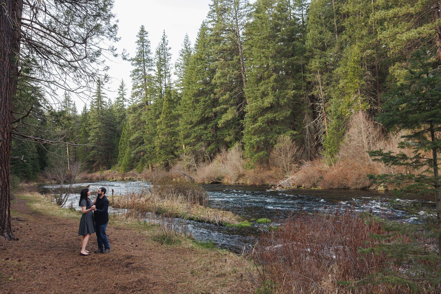 Couple embracing on a forest trail beside a flowing river surrounded by tall pine trees.