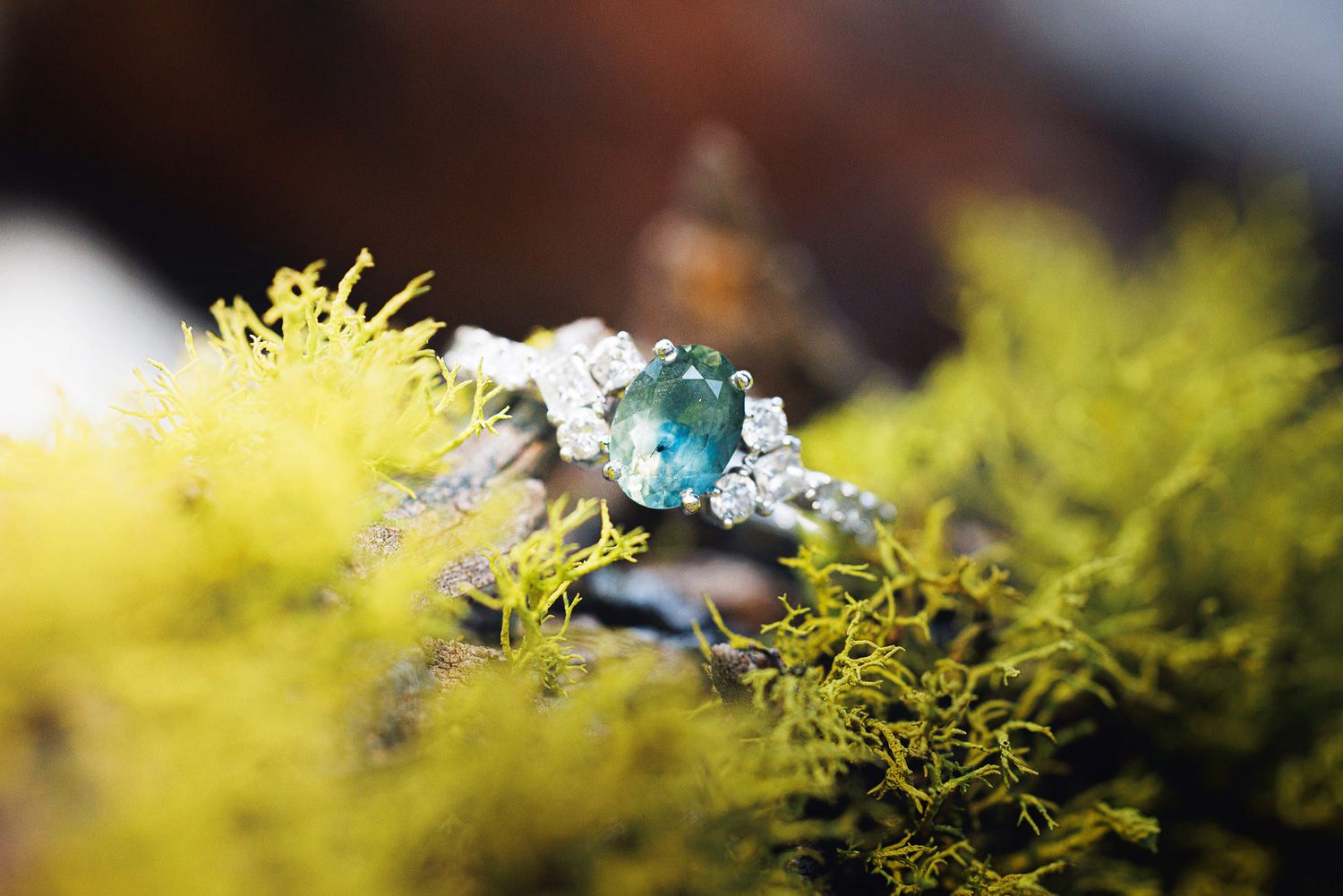 Silver ring with teal gemstone and diamond accents resting on vibrant green moss in a natural outdoor setting.