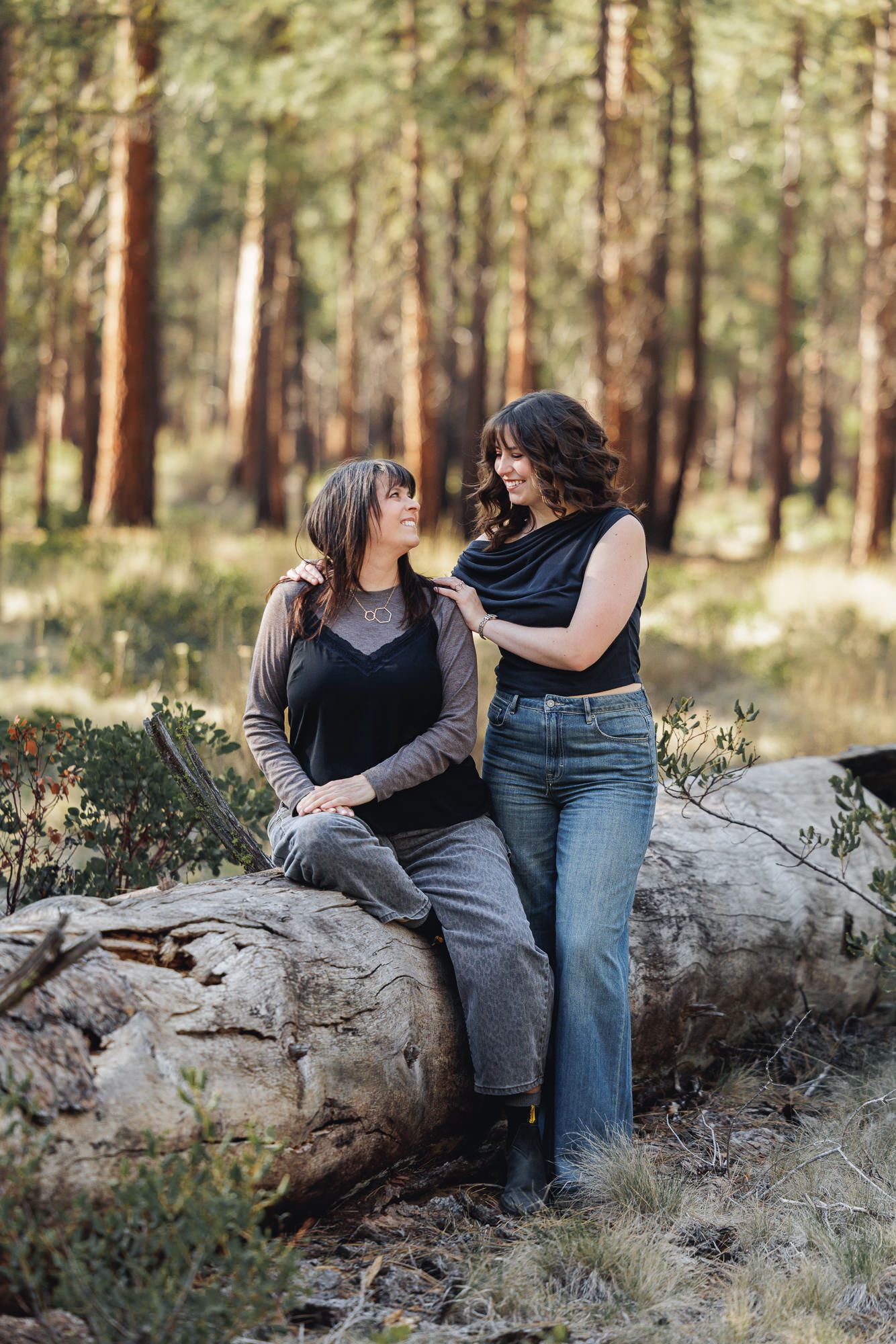 Two women share a warm moment on a fallen log in a sunlit pine forest, one seated and one standing beside her.