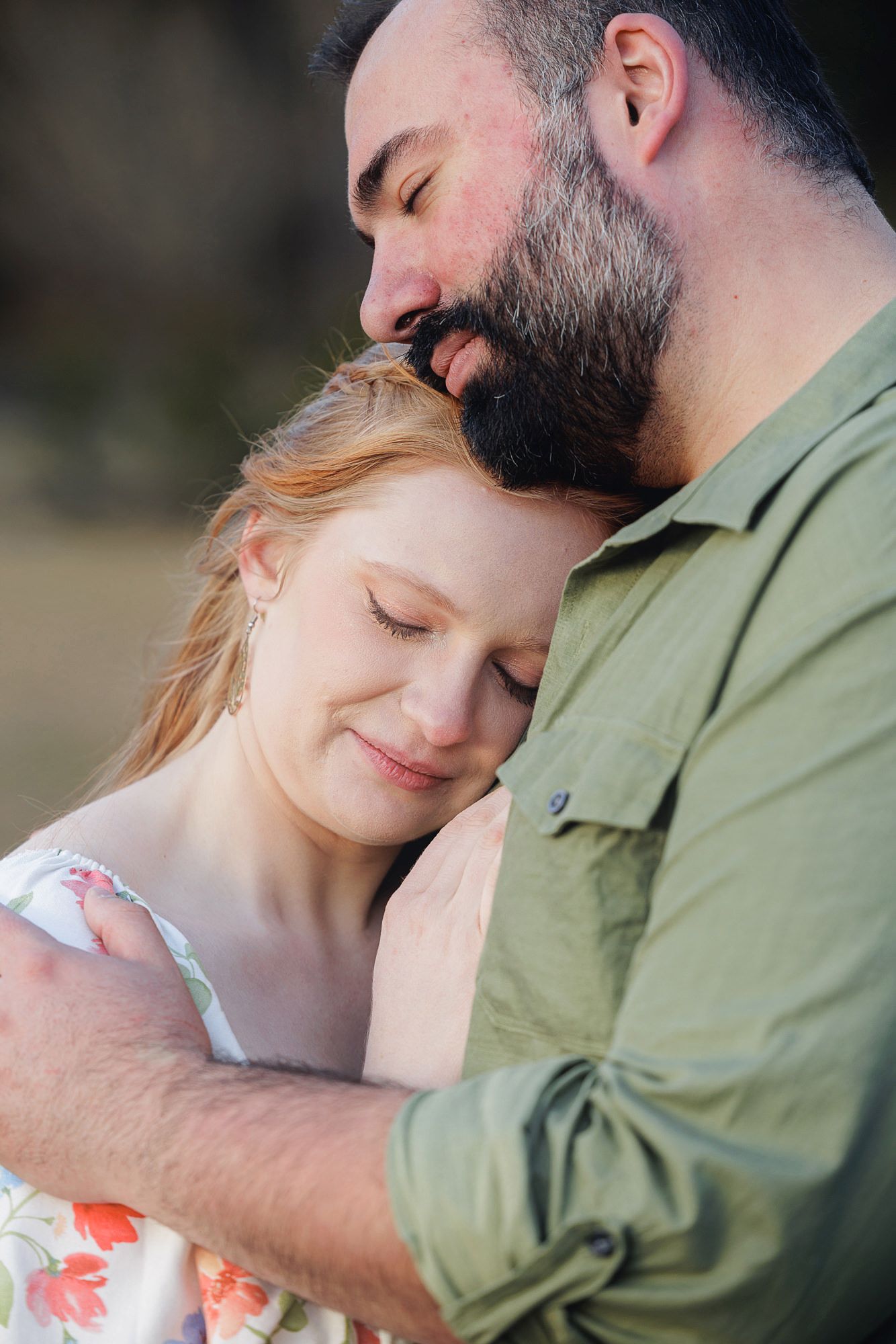 A bearded man in a green shirt tenderly embraces a smiling redhead woman in a floral dress outdoors.