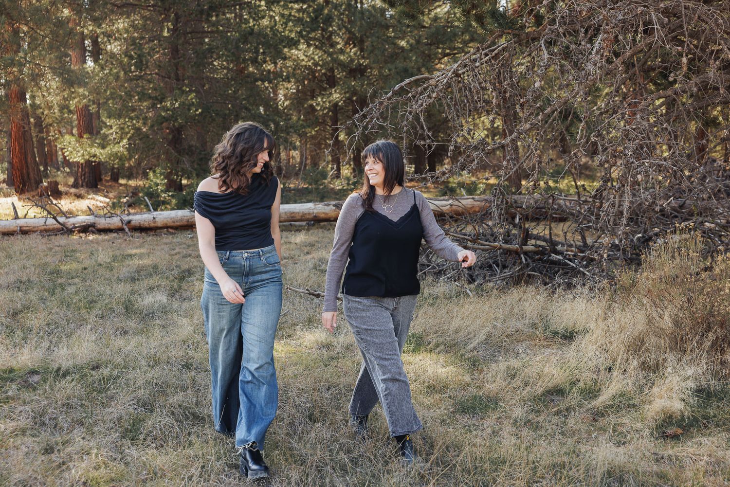 Two women walking outdoors in a forest meadow, wearing casual outfits of jeans and tops, surrounded by trees.