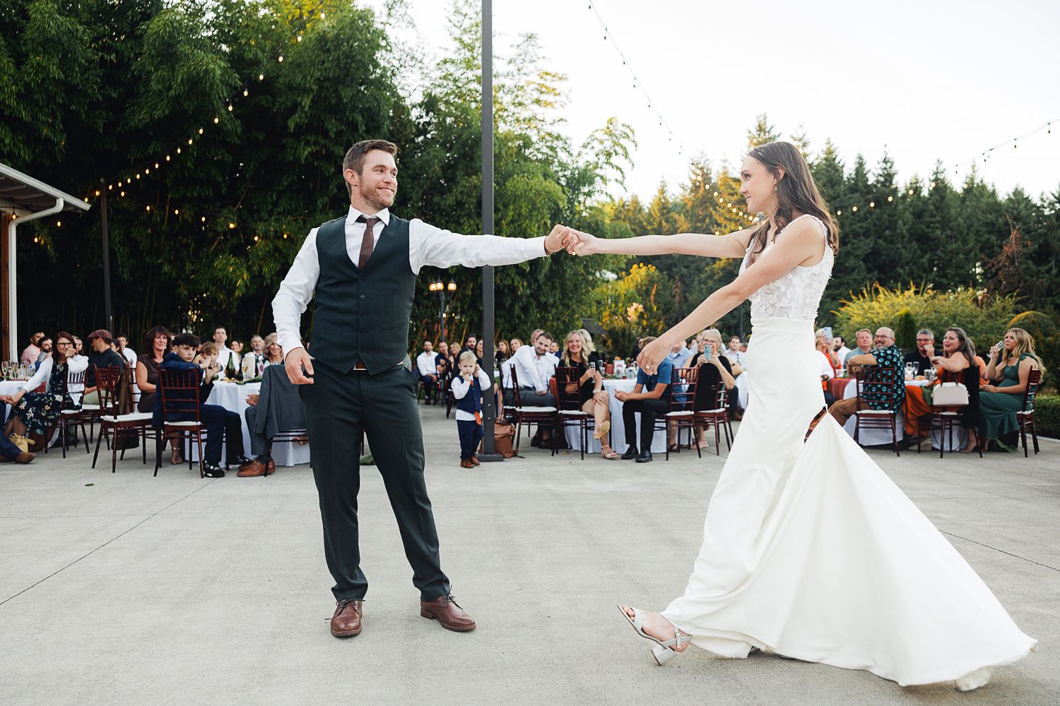 Couple performs a playful dance sequence at an outdoor wedding reception with guests watching in the background.