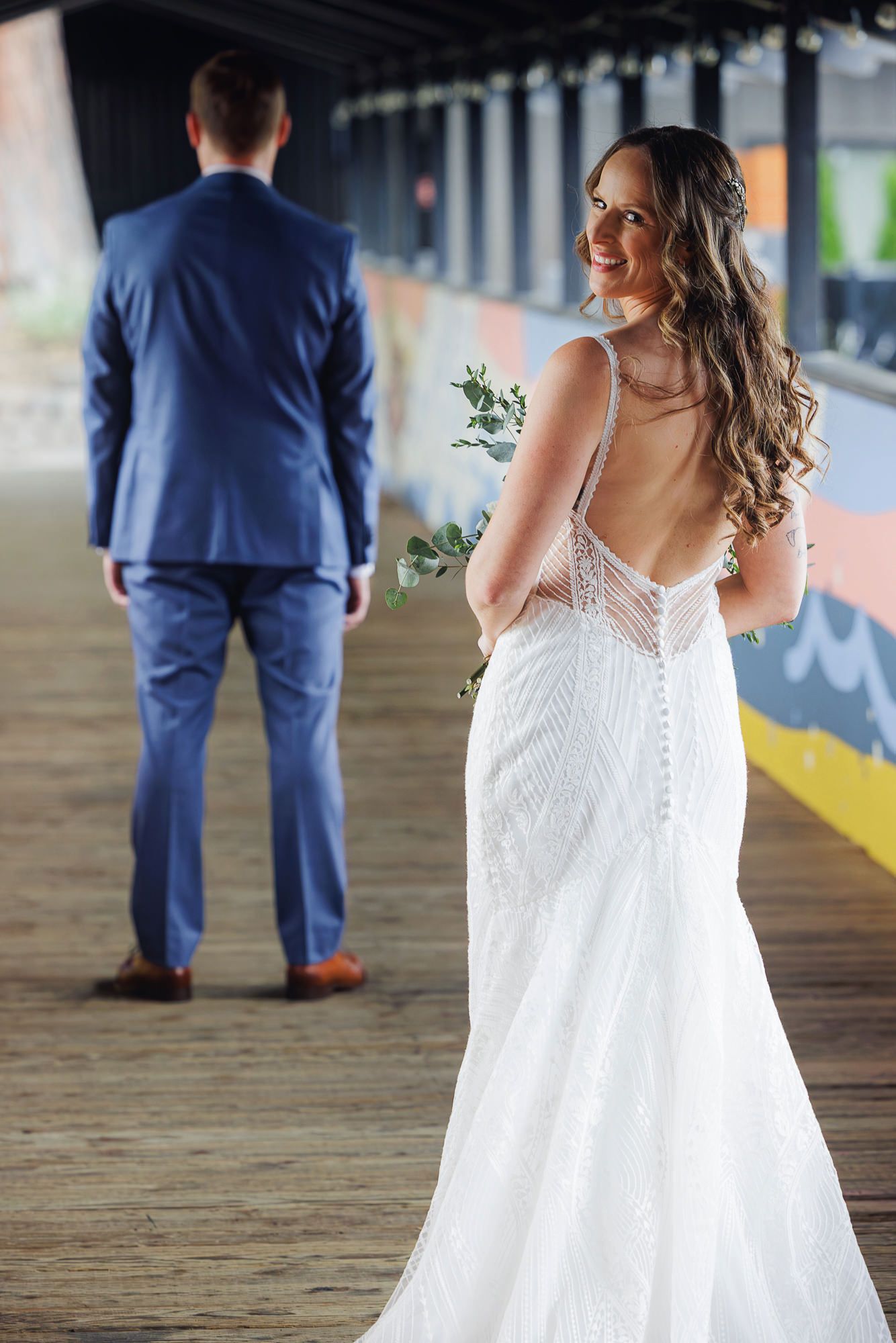 A bride in a white lace dress and groom in navy suit walk separately along a covered outdoor walkway.