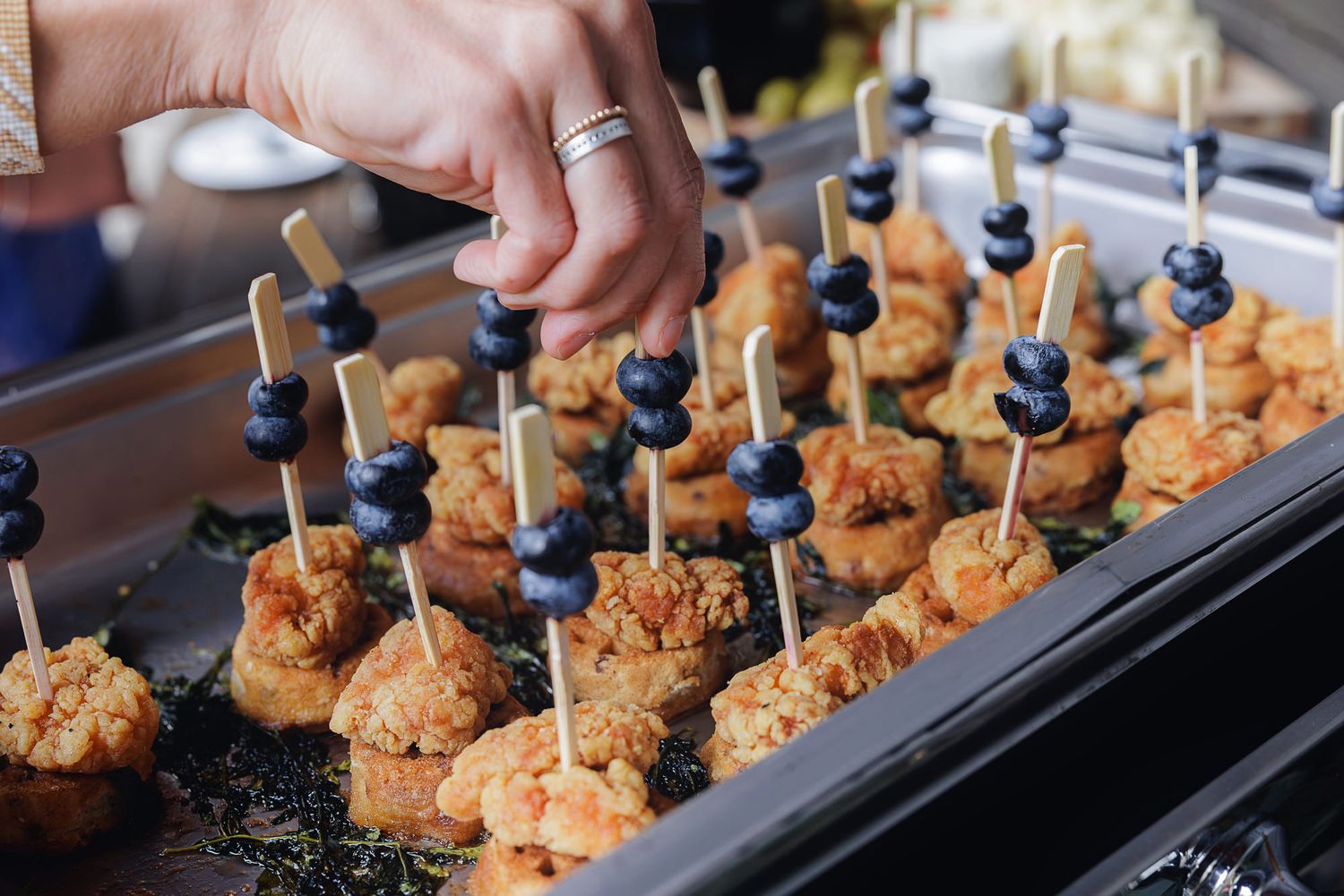 Close-up of hands placing crispy fried shrimp appetizers with toothpicks on a dark serving platter.