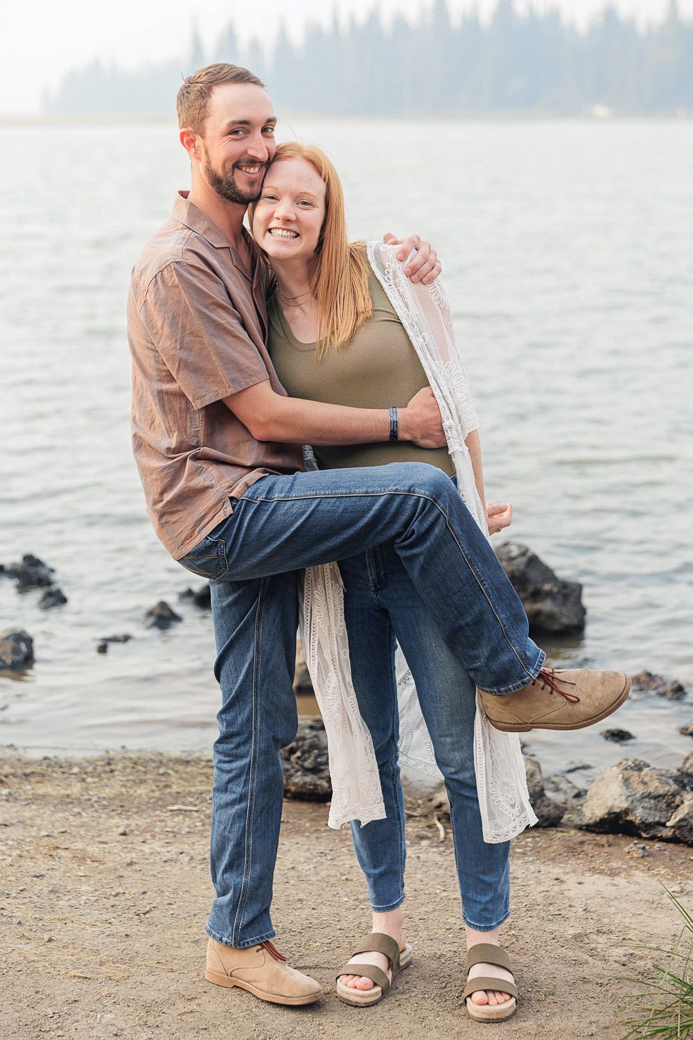 A romantic couple sits together on driftwood by a misty lake shore.