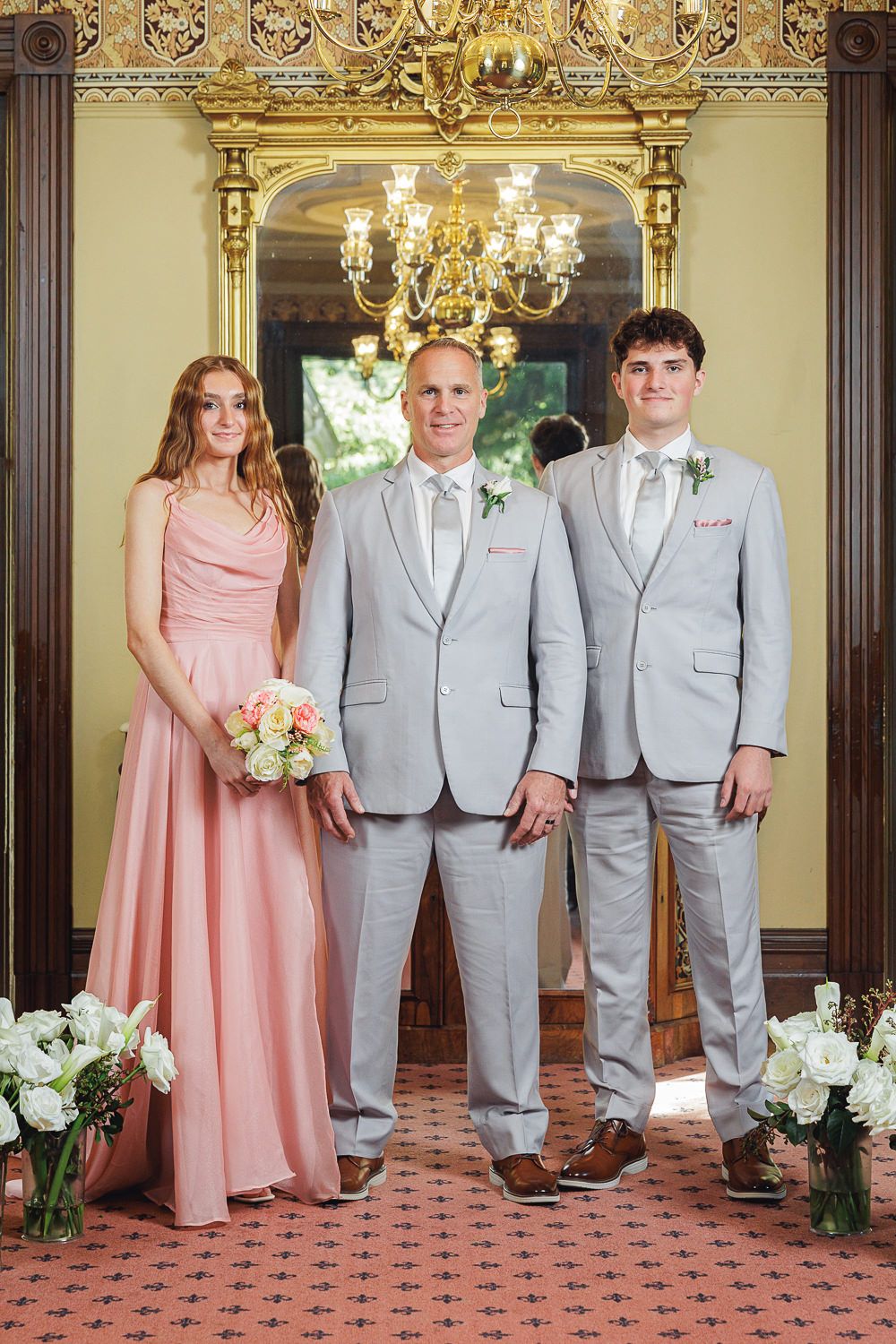 Three people in formal attire stand in front of an ornate doorway with a chandelier and floral arrangements.