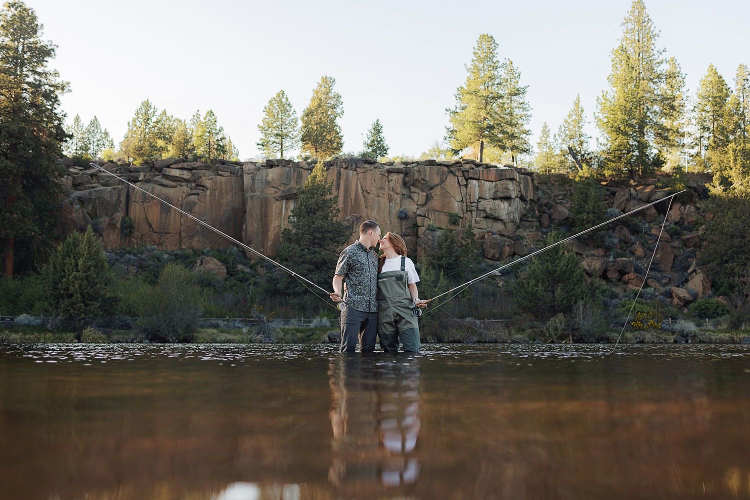 Two people stand together in shallow water near rock cliffs with pine trees against a serene natural backdrop.