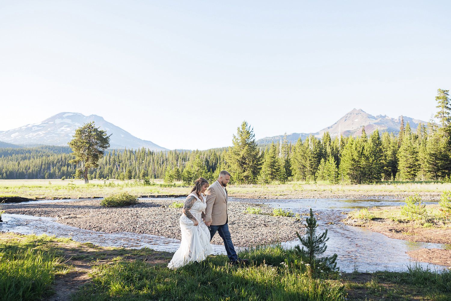 Newlywed couple walks near a scenic mountain lake surrounded by evergreen forest and distant peaks.
