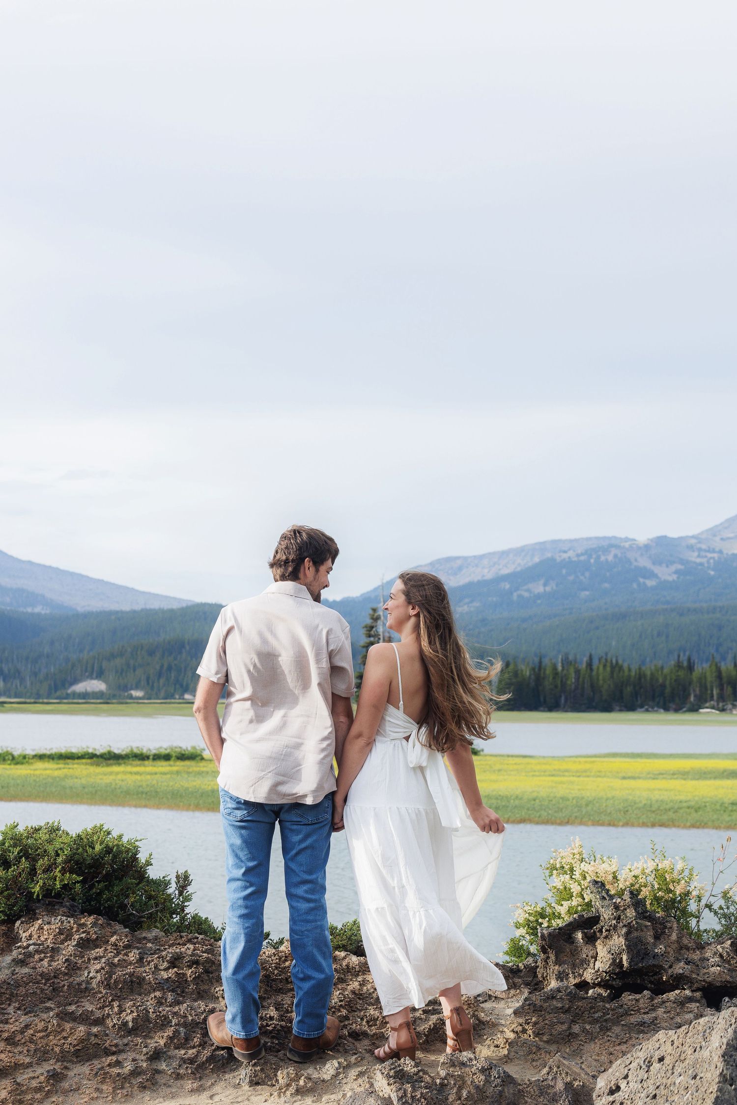 Couple in white dress and casual attire share intimate moment near mountain lake with scenic mountain backdrop.