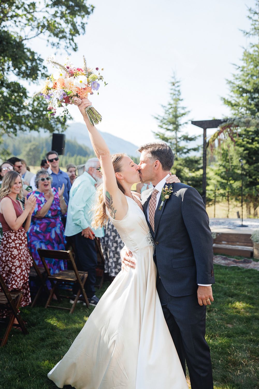 Wedding guests celebrate and toast during an outdoor reception with mountains and evergreen trees in the background.