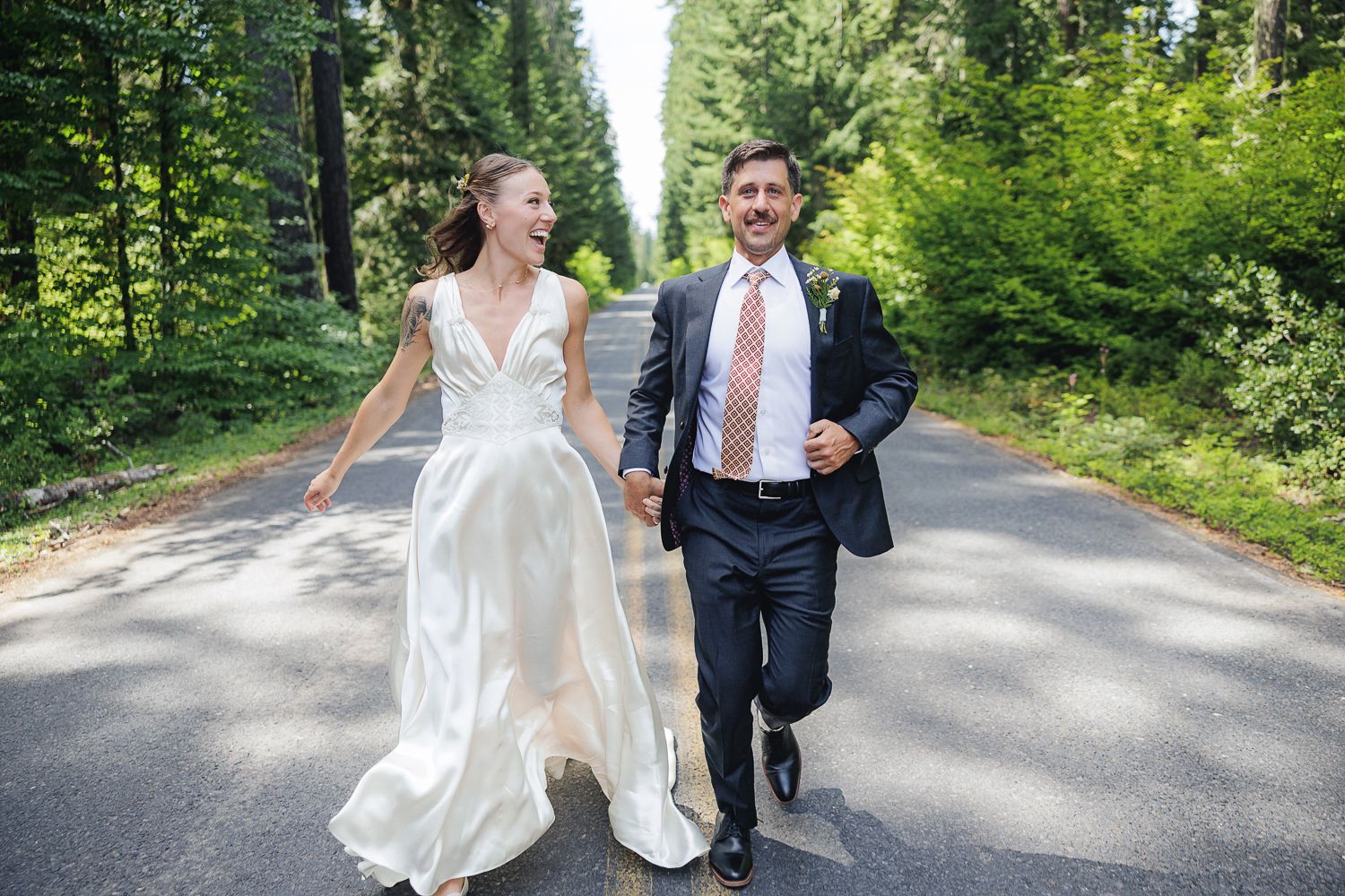 Happy couple in wedding attire running playfully together down a tree-lined road on their wedding day.