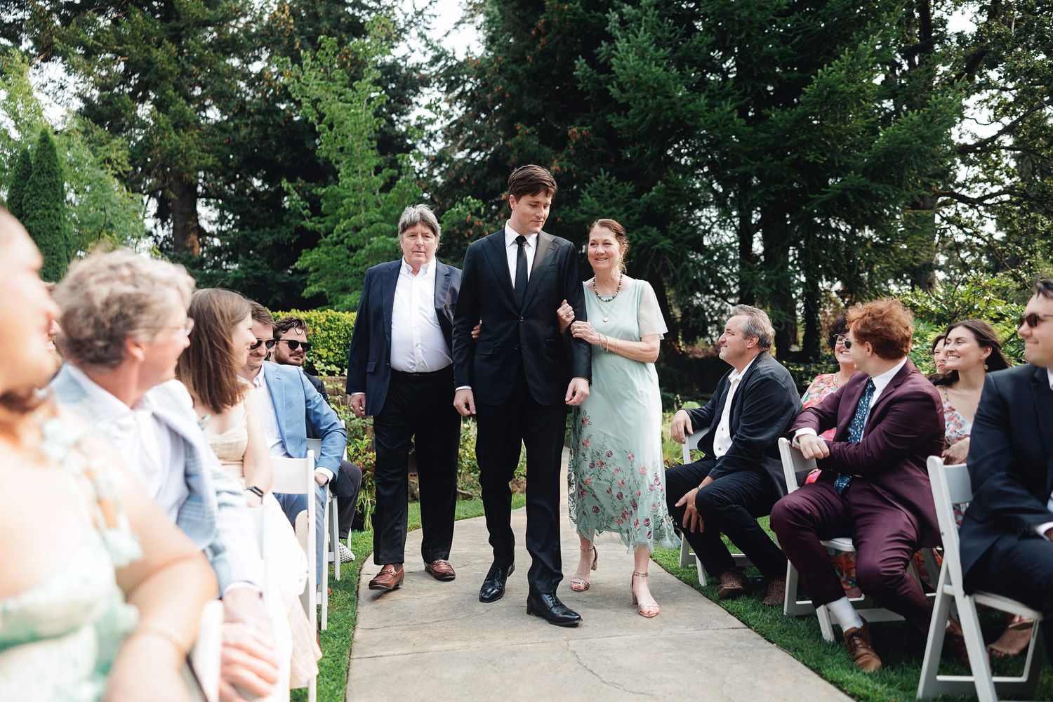A young man in a black suit walks down a wedding aisle escorting a woman in a floral dress outdoors.