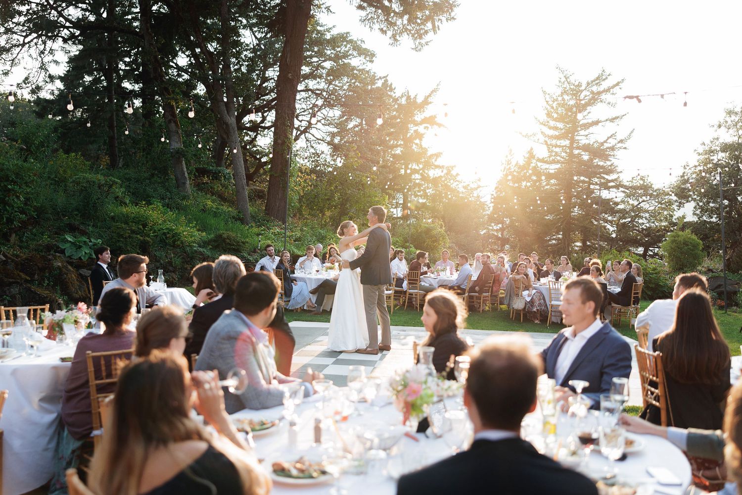 Couple shares first dance at golden hour outdoor wedding reception with guests seated at elegant tables.